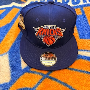 May include: A royal blue New York Knicks snapback cap featuring an embroidered team logo on the front with orange and white accents. The hat includes a circular 9FIFTY sticker on the flat brim and a commemorative patch on the side panel.
