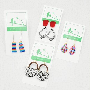 May include: Four pairs of earrings displayed on white cards. The earrings feature various designs, including red squares with black and white geometric shapes, colorful confetti patterns, and rainbow stripes. The cards have a green logo and the text "J. Byrd creative."