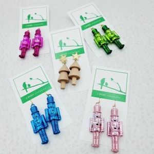 Puede incluir: Varios pares de pendientes se muestran en tarjetas blancas. Los pendientes incluyen cascanueces rosas, azules y verdes, árboles de Navidad de madera y detalles de estrellas doradas. Las tarjetas tienen un logotipo verde con el texto "J. BYRD creative."