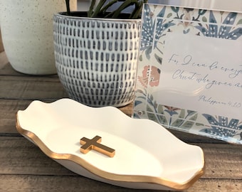 May include: A white ceramic dish with a gold rim and a gold cross in the center. The dish has a scalloped edge. The background includes a framed print with floral designs and a potted plant.