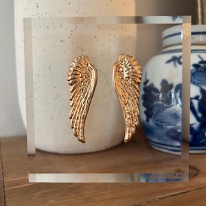 May include: A clear acrylic block featuring two gold-colored angel wings. The wings are detailed with textured feathers. The block is displayed on a wooden surface, with a white ceramic vase and a blue and white ceramic jar in the background.