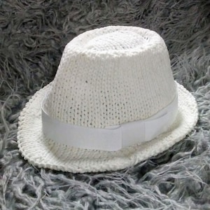 May include: A white fedora hat with a knitted texture. The hat features a white ribbon band around the crown. The brim is slightly upturned. The hat is sitting on a gray, textured surface.