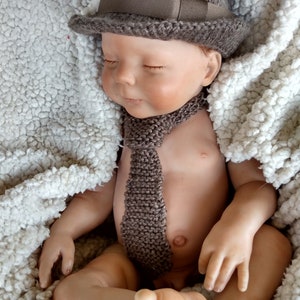 Peut inclure: Un b&eacute;b&eacute; poup&eacute;e endormi portant un chapeau en tricot marron avec un accent de plume et une cravate en tricot marron.