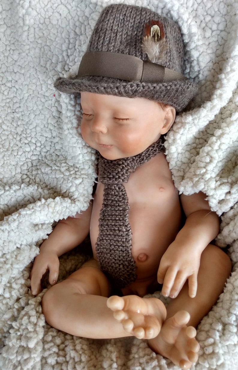 Peut inclure: Un b&eacute;b&eacute; poup&eacute;e endormi portant un chapeau en tricot marron avec un accent de plume et une cravate en tricot marron.