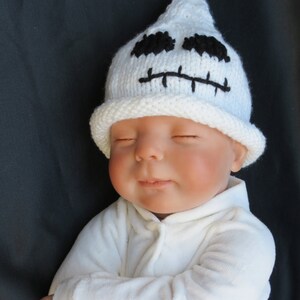 May include: A white knitted ghost hat with black embroidered eyes and mouth on a baby wearing a white onesie.