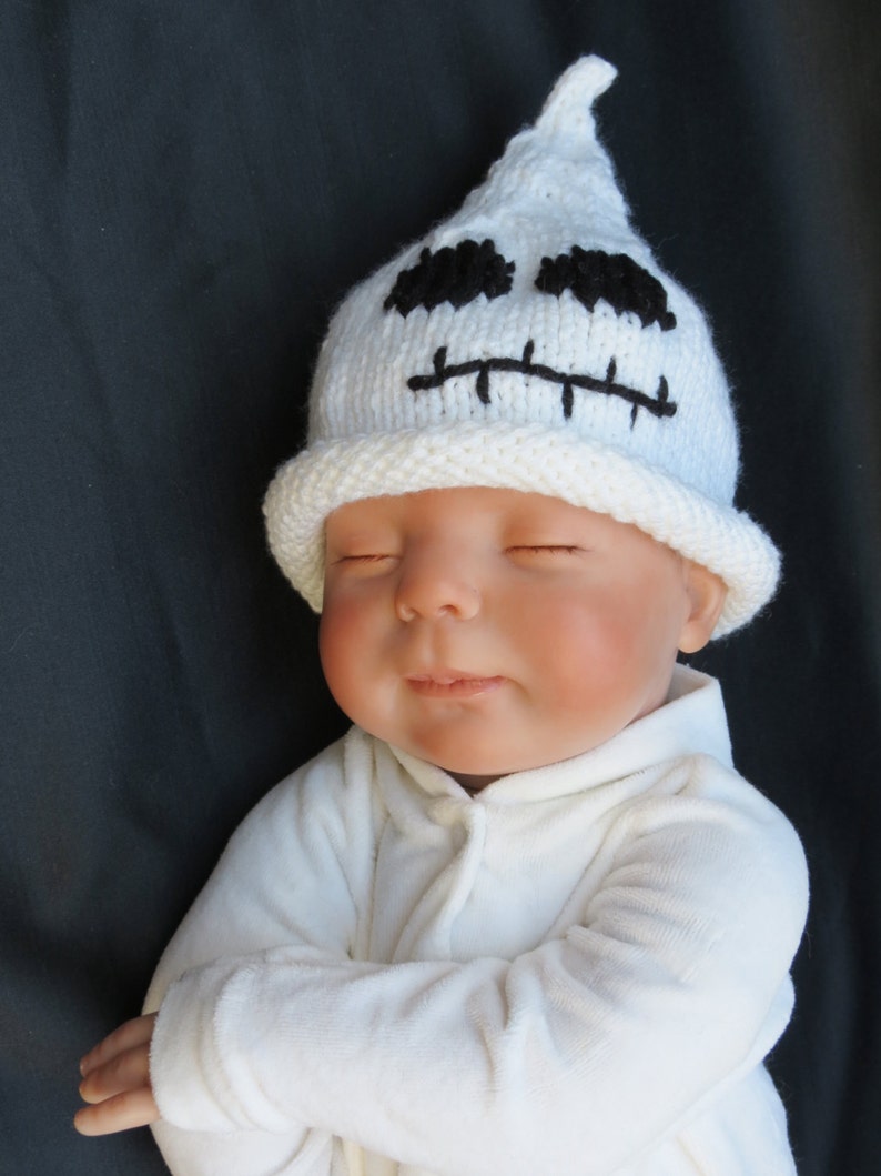 May include: A white knitted ghost hat with black embroidered eyes and mouth on a baby wearing a white onesie.