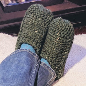 May include: A pair of green knitted slippers with a flecked texture. The slippers are worn by a person sitting in front of a fireplace.