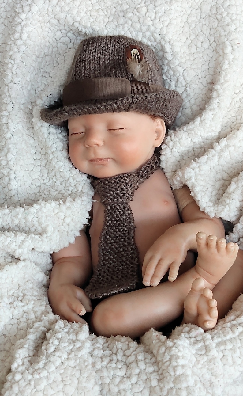 Peut inclure: Un b&eacute;b&eacute; poup&eacute;e endormi portant un chapeau en tricot marron avec un ruban marron et une cravate en tricot marron. La poup&eacute;e est allong&eacute;e sur une couverture moelleuse blanche.