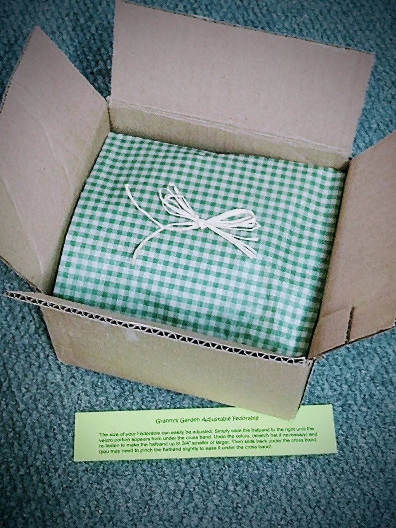 Peut inclure: Une bo&icirc;te en carton avec un papier de soie &agrave; carreaux verts et blancs. La bo&icirc;te est ouverte et le papier de soie est visible. Une petite carte verte et blanche avec le texte "Granny's Garden Adjustable Fedorable" est visible dans le coin inf&eacute;rieur droit de l'image. Le texte sur la carte indique : "La taille de votre Fedorable peut &ecirc;tre facilement ajust&eacute;e. Faites simplement glisser le bandeau vers la droite jusqu'&agrave; ce que la partie velcro apparaisse sous le bandeau crois&eacute;. D&eacute;faites le velcro, (&eacute;tirez le chapeau si n&eacute;cessaire) et refixez-le pour rendre le bandeau jusqu'&agrave; 3/4" plus petit ou plus grand. Ensuite, glissez-le &agrave; nouveau sous le bandeau crois&eacute; (vous devrez peut-&ecirc;tre pincer l&eacute;g&egrave;rement le bandeau pour le faire passer sous le bandeau crois&eacute;)"