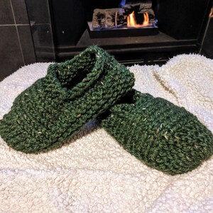May include: A pair of green knitted slippers with a flecked texture. The slippers are on a white fluffy surface.