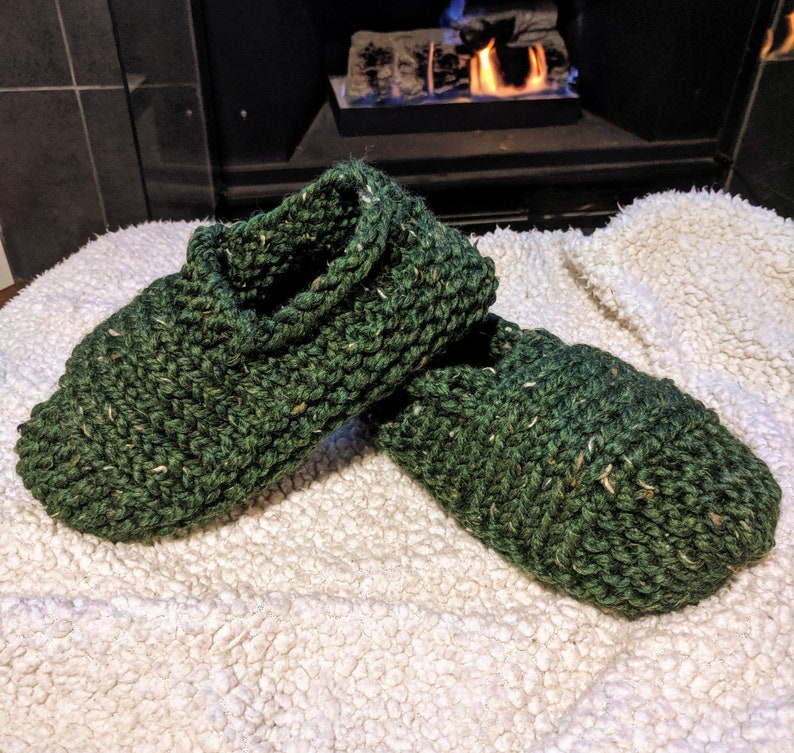 May include: A pair of green knitted slippers with a flecked texture. The slippers are on a white fluffy surface.