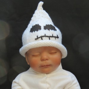 May include: A white knitted ghost hat with black embroidered eyes and mouth. The hat is worn by a baby with closed eyes.
