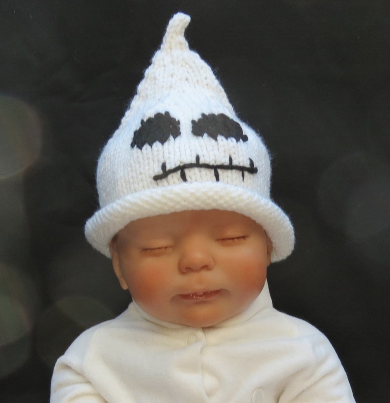 May include: A white knitted ghost hat with black embroidered eyes and mouth. The hat is worn by a baby with closed eyes.