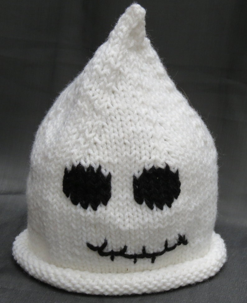 May include: White knitted hat with a ghost face design. The ghost has black eyes and a stitched mouth.