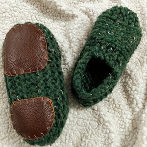 May include: A pair of green knitted slippers with brown leather patches. The slippers are on a white fluffy surface.