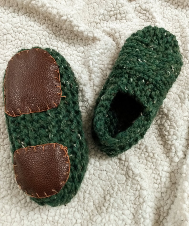 May include: A pair of green knitted slippers with brown leather patches. The slippers are on a white fluffy surface.