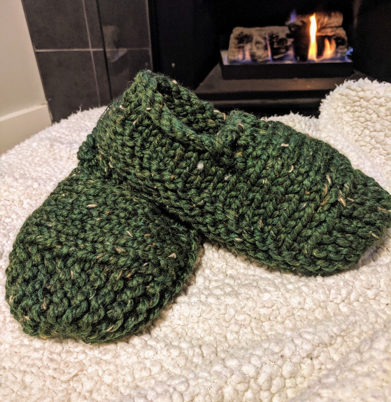 May include: A pair of green knitted slippers with a flecked texture. The slippers are sitting on a white fluffy surface.