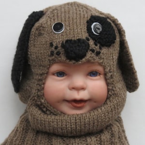 May include: A brown knitted baby hat with a dog face design. The hat has black ears, a black nose, and two black eyes with white buttons. The hat is worn by a baby with blue eyes.