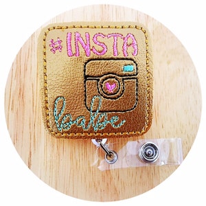 May include: Gold-colored square badge holder with a black and white camera design, pink hashtag "#INSTA babe" text, and a clear retractable badge reel.