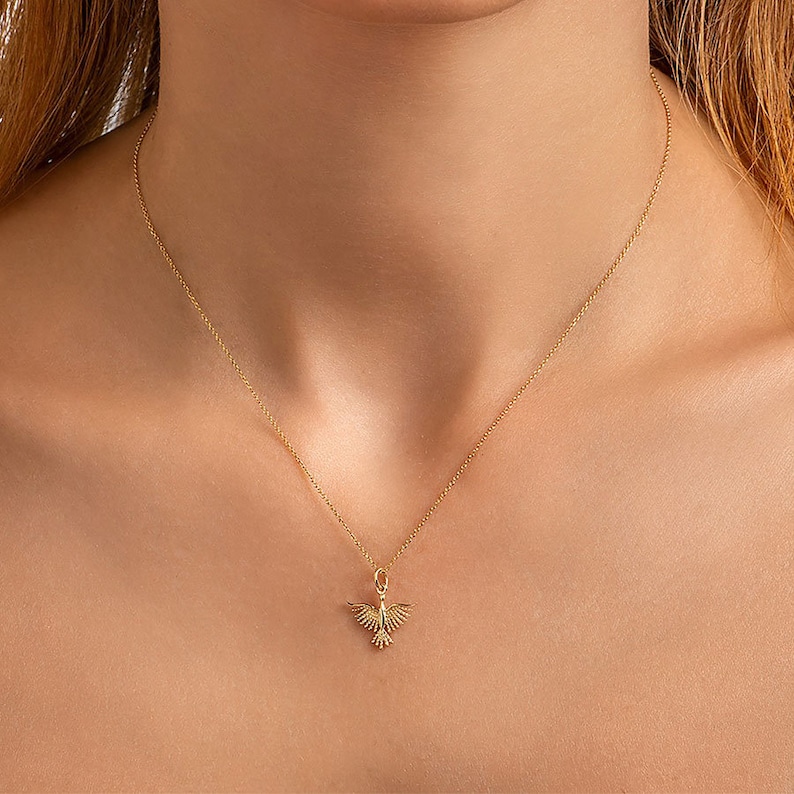 May include: A gold chain necklace with a delicate bird pendant. The bird is facing forward with its wings spread out.