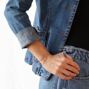 May include: A gold chain bracelet with a small puzzle piece charm. The bracelet is worn on a person's wrist, and the person is wearing a denim jacket and jeans.