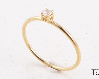 Op de afbeelding: Een delicate gouden ring met een enkele, kleine, heldere edelsteen gezet in een eenvoudige, vierkante kraalbezetting.