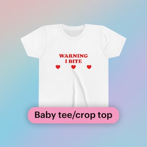 May include: White baby tee or crop top with the text "WARNING I BITE" in red, along with three red heart symbols. The shirt has a crew neck and short sleeves. The bottom of the image has the text "Baby tee/crop top" in a pink rectangle.