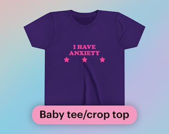 Może przedstawiać: Fioletowa koszulka dziecięca lub crop top z napisem "I HAVE ANXIETY" w różowych literach i trzema różowymi gwiazdkami.