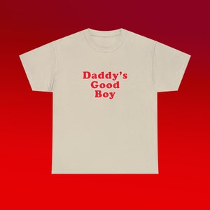 May include: A beige t-shirt with the text "Daddy's Good Boy" printed in red.