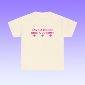 Puede incluir: Una camiseta beige con el texto "SAVE A HORSE RIDE A COWBOY" en letras rosas y tres estrellas rosas debajo del texto.