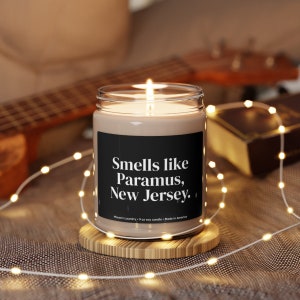 May include: A brown soy candle with a black label that reads "Smells like Paramus, New Jersey." The candle is lit and sitting on a wooden coaster.