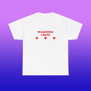 Puede incluir: Camiseta blanca con el texto "WARNING I BITE" en letras rojas y tres corazones rojos.