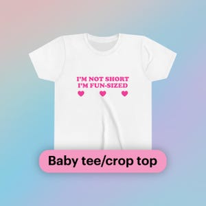 Pode incluir: Camiseta branca para bebê ou top cropped com texto rosa que diz "Não sou baixa, sou do tamanho divertido" com três corações rosas abaixo do texto.