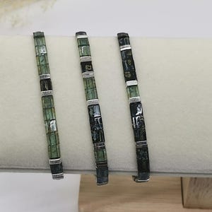 Peut inclure: Trois bracelets de perles avec des perles carrées vertes et noires, séparées par des accents métalliques argentés. Les bracelets sont présentés sur une surface blanche.