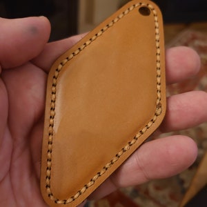 May include: A brown leather key fob with a diamond shape and a stitched edge. The fob has a hole at the top for attaching a key ring.