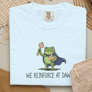 Könnte beinhalten: Ein hellblaues T-Shirt mit einer Grafik eines grünen Frosches in einem blauen Umhang, der einen goldenen Schlüssel und drei gelbe Sterne hält. Der Text "WE REINFORCE AT DAWN" ist unter dem Frosch gedruckt. Das Shirt hat ein Comfort Colors Etikett.