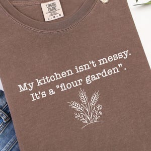 Op de afbeelding: Een bruin t-shirt met de tekst "My kitchen isn't messy. It's a 'flour garden'" en een tarwe- en bloemenafbeelding. Het shirt is opgevouwen met een spijkerbroek en een gouden ketting.