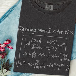 Puede incluir: Una camiseta negra con el texto "Retiring once I solve this" y ecuaciones matemáticas blancas. La camiseta está sobre unos vaqueros azules, un sombrero rosa y un ramo de flores rosas.