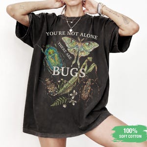 May include: A charcoal black short-sleeve t-shirt featuring a graphic print of various insects and plants. The text reads "YOU'RE NOT ALONE THERE ARE BUGS". The shirt includes a green rectangular graphic in the corner with the text "100% SOFT COTTON".