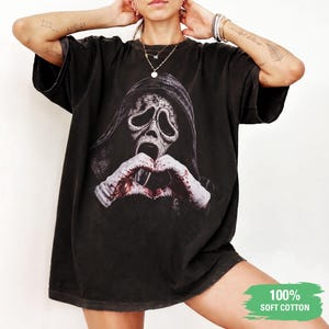 Pode incluir: Uma t-shirt preta de manga curta com uma estampa gráfica de uma figura encapuzada com máscara branca e mãos manchadas de sangue formando um coração. O tecido tem uma aparência vintage desbotada. Um elemento gráfico verde indica 100% SOFT COTTON.