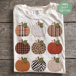 Peut inclure: Un t-shirt couleur crème arborant une grille de neuf citrouilles aux motifs variés, incluant des carreaux, des pois, des rayures, des imprimés léopard et zèbre. Chaque citrouille a une feuille verte. Une étiquette indique 100% SOFT COTTON.