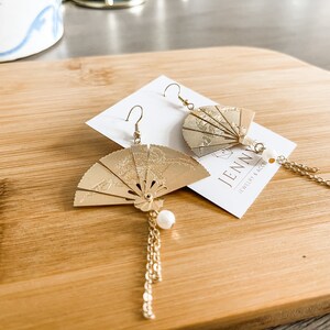 May include: Gold fan-shaped earrings with a pearl charm and a gold chain dangle. The earrings are on a wooden surface with a business card that says "JENN Jewelry & Accessories".