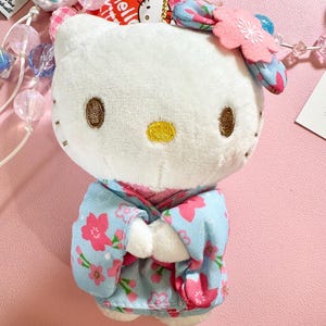 May include: A white plush Hello Kitty doll wearing a light blue kimono with pink floral patterns. The doll has brown embroidered eyes and a yellow nose. A pink flower hair accessory and a gold keychain are attached.