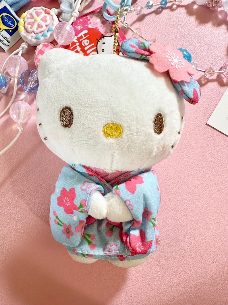 May include: A white plush Hello Kitty doll wearing a light blue kimono with pink floral patterns. The doll has brown embroidered eyes and a yellow nose. A pink flower hair accessory and a gold keychain are attached.