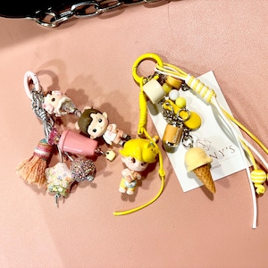 May include: Two keychains with charms. One keychain has a pink and silver tassel, a pink cup, and small figurines. The other keychain has yellow accents, a yellow ice cream cone, and a small can charm.