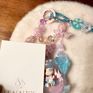 May include: A decorative keychain with a pink clasp, adorned with pastel-colored beads, flowers, and a small figurine of a character with a bunny. The keychain has a light blue and pink color scheme, with the text "JENNY'S CORNER" on a white card.