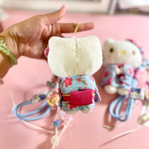 May include: A plush Hello Kitty doll wearing a blue kimono with a floral pattern and a pink obi. The doll has a white head and is held by a hand. A second doll is slightly out of focus in the background.