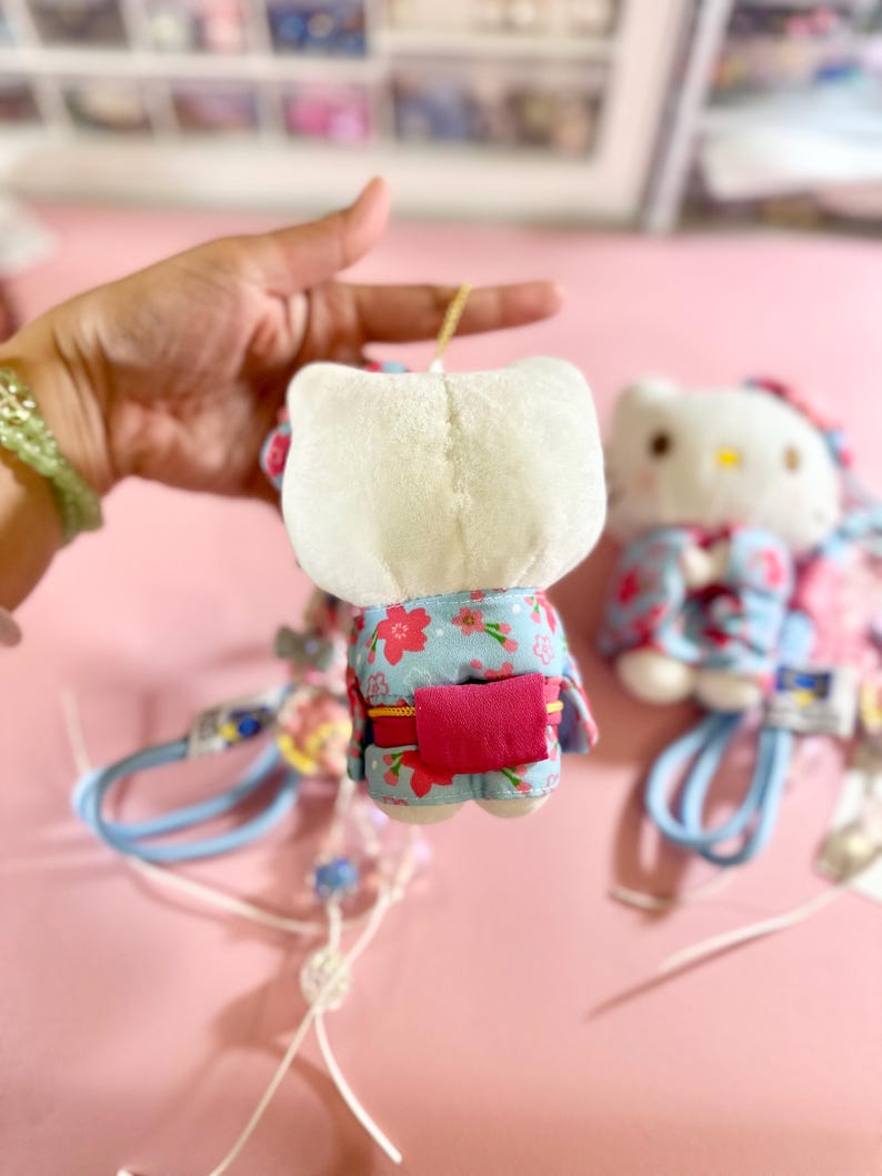 May include: A plush Hello Kitty doll wearing a blue kimono with a floral pattern and a pink obi. The doll has a white head and is held by a hand. A second doll is slightly out of focus in the background.