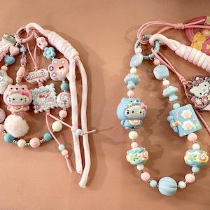 May include: Two decorative keychains featuring Hello Kitty charms, beads, and ribbons. The keychains have pink, blue, and white accents. One keychain has a white pom-pom. The charms include Hello Kitty figures and other decorative shapes.