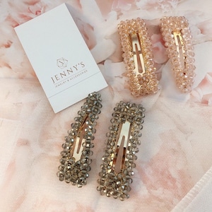 May include: Four beaded hair clips in gold and gray tones. Two rectangular clips are a light peach color, and two are gray. The clips are embellished with small, round beads. The brand name "Jenny's Jewelry & Accessories" is visible on a white card.